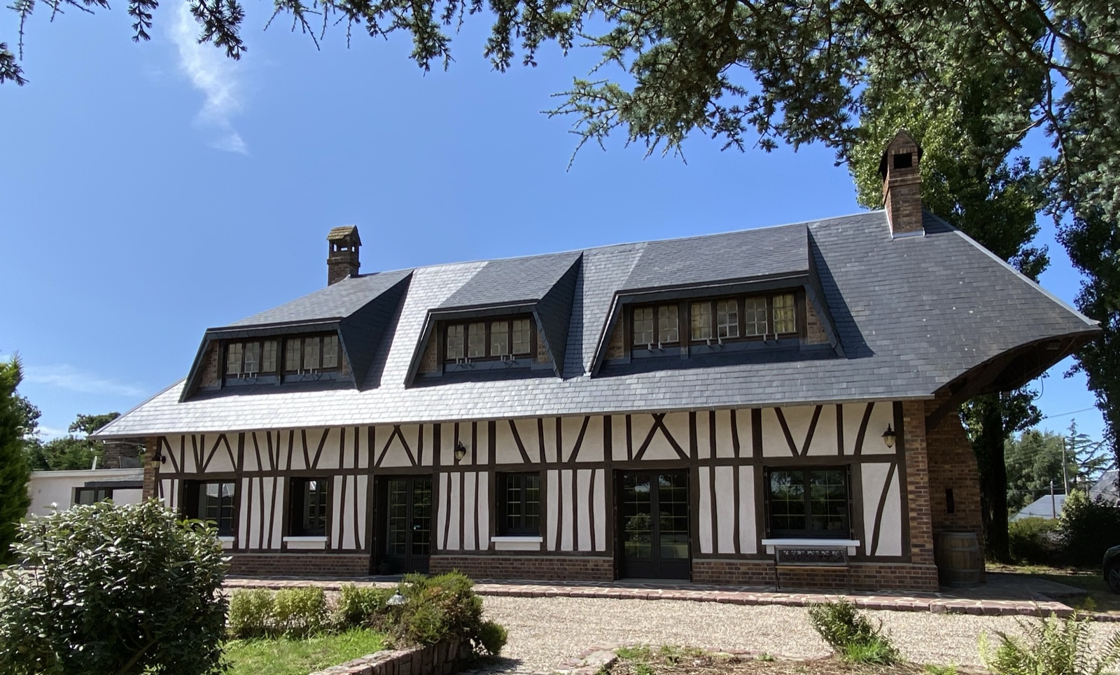 Long elevation of the half-timbered façade with three slate dormers, framed by an overhanging cedar branch